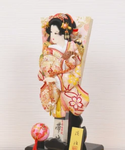 Hagoita paddle with elegant kimono-clad female figure, with gold and red embroidery, displayed on a black lacquered base and display case with decorative ball. Wholesale by Fujiki Crafts.