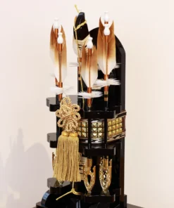 Japanese Hamaya arrow display with gold cord and tassel in black lacquer case, set against gold background with feathered arrows and symbolic ornaments. Wholesale by Fujiki Crafts.