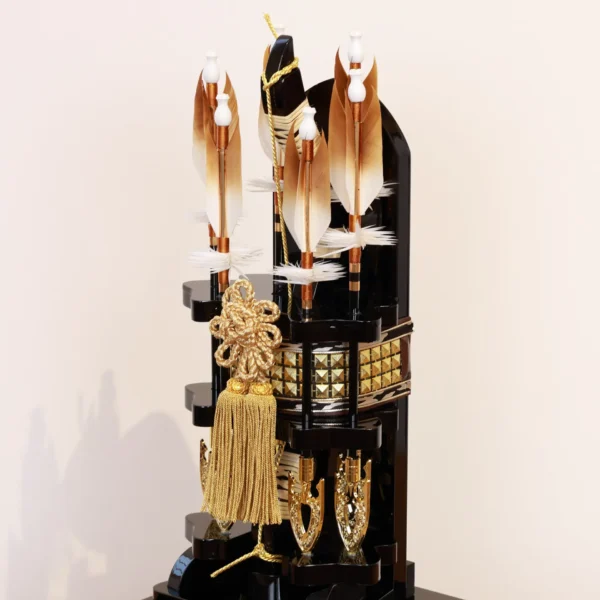 Japanese Hamaya arrow display with gold cord and tassel in black lacquer case, set against gold background with feathered arrows and symbolic ornaments. Wholesale by Fujiki Crafts.