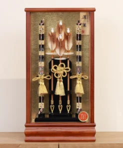 Japanese Hamaya arrow display with gold cord tassels and feathered arrows in a wooden frame case, set against a gold textured backdrop. Wholesale by Fujiki Crafts.
