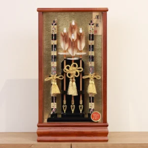 Japanese Hamaya arrow display with gold cord tassels and feathered arrows in a wooden frame case, set against a gold textured backdrop. Wholesale by Fujiki Crafts.