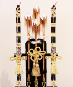 Japanese Hamaya arrow display with gold cord tassels and feathered arrows in a wooden frame case, set against a gold textured backdrop. Wholesale by Fujiki Crafts.