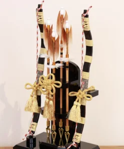 Japanese Hamaya arrow display with gold cord tassels and feathered arrows in a wooden frame case, set against a gold textured backdrop. Wholesale by Fujiki Crafts.