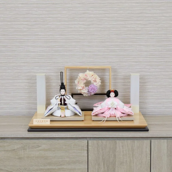 Modern Japanese Hina doll set with white and pink kimono dolls on wooden platform, decorated with pastel floral wreath and minimalist lighting stands. Wholesale by Fujiki Crafts.