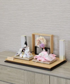Modern Japanese Hina doll set with white and pink kimono dolls on wooden platform, decorated with pastel floral wreath and minimalist lighting stands. Wholesale by Fujiki Crafts.