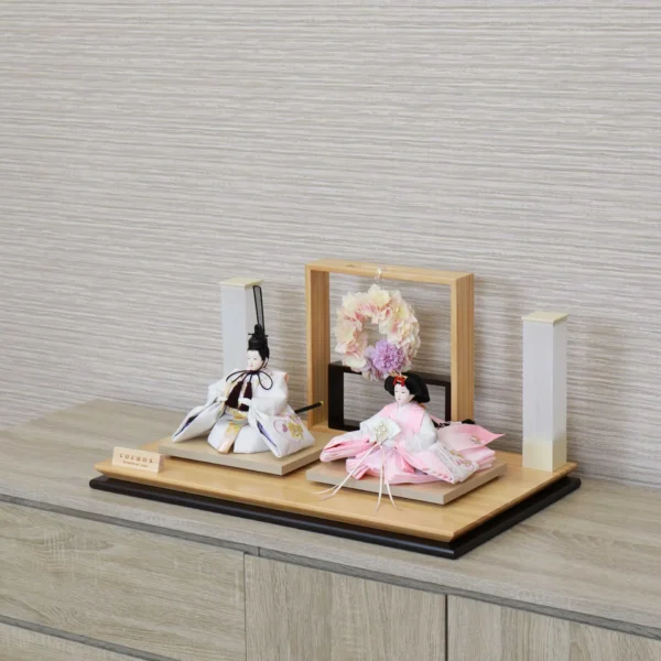 Modern Japanese Hina doll set with white and pink kimono dolls on wooden platform, decorated with pastel floral wreath and minimalist lighting stands. Wholesale by Fujiki Crafts.
