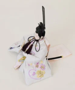 Emperor Hina doll in white patterned kimono with gold and purple accents, holding a ceremonial baton. Wholesale by Fujiki Crafts.
