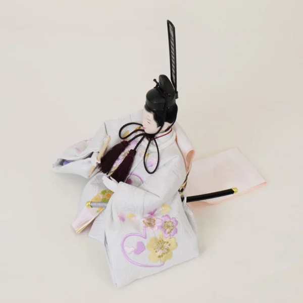 Emperor Hina doll in white patterned kimono with gold and purple accents, holding a ceremonial baton. Wholesale by Fujiki Crafts.