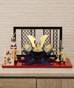 Japanese Kabuto helmet display with gold crest and silk thread navy strap on red lacquered base, featuring folding screen, miniature sword, and decorative arrows. Wholesale by Fujiki Crafts.