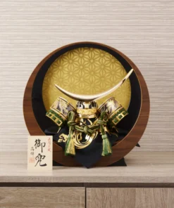 Japanese Kabuto helmet with gold crescent crest and green cord accents, set in a round wooden frame with gold patterned background. Wholesale by Fujiki Crafts.