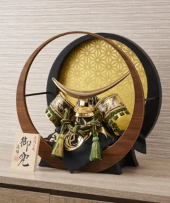Japanese Kabuto helmet with gold crescent crest and green cord accents, set in a round wooden frame with gold patterned background. Wholesale by Fujiki Crafts.