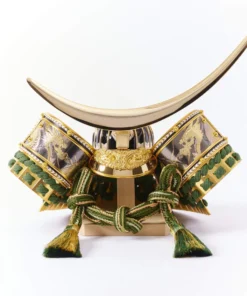 Japanese Kabuto helmet with gold crescent crest and green cord accents, set in a round wooden frame with gold patterned background. Wholesale by Fujiki Crafts.