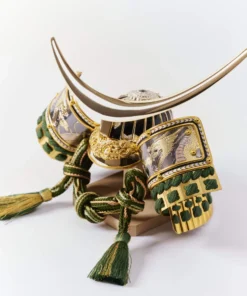 Japanese Kabuto helmet with gold crescent crest and green cord accents, set in a round wooden frame with gold patterned background. Wholesale by Fujiki Crafts.