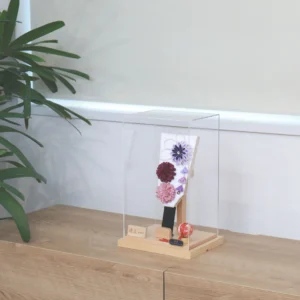 Handcrafted Japanese Hagoita paddle in display case with soft pink and purple flowers, set on wooden base with Yuubi nameplate. Wholesale by Fujiki Crafts.