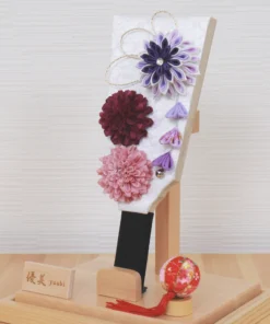 Handcrafted Japanese Hagoita paddle in display case with soft pink and purple flowers, set on wooden base with Yuubi nameplate. Wholesale by Fujiki Crafts.