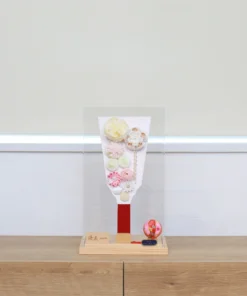 Handcrafted Japanese Hagoita paddle in pastel tones, decorated with white and pink tsumami flowers, presented in a clear acrylic display case with red handle and wooden base. Wholesale by Fujiki Crafts.