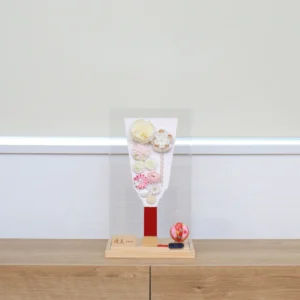 Handcrafted Japanese Hagoita paddle in pastel tones, decorated with white and pink tsumami flowers, presented in a clear acrylic display case with red handle and wooden base. Wholesale by Fujiki Crafts.