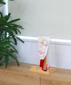 Handcrafted Japanese Hagoita paddle in pastel tones, decorated with white and pink tsumami flowers, presented in a clear acrylic display case with red handle and wooden base. Wholesale by Fujiki Crafts.