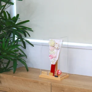 Handcrafted Japanese Hagoita paddle in pastel tones, decorated with white and pink tsumami flowers, presented in a clear acrylic display case with red handle and wooden base. Wholesale by Fujiki Crafts.