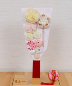 Handcrafted Japanese Hagoita paddle in pastel tones, decorated with white and pink tsumami flowers, presented in a clear acrylic display case with red handle and wooden base. Wholesale by Fujiki Crafts.