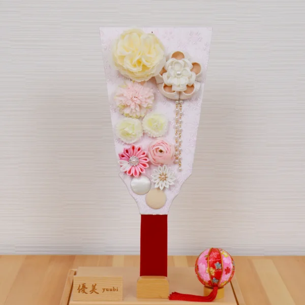 Handcrafted Japanese Hagoita paddle in pastel tones, decorated with white and pink tsumami flowers, presented in a clear acrylic display case with red handle and wooden base. Wholesale by Fujiki Crafts.