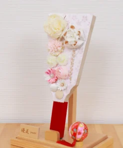 Handcrafted Japanese Hagoita paddle in pastel tones, decorated with white and pink tsumami flowers, presented in a clear acrylic display case with red handle and wooden base. Wholesale by Fujiki Crafts.