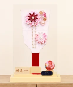Handcrafted Japanese Hagoita paddle in soft pastel pink tones, adorned with white and pink tsumami kanzashi flowers, pearl accents, and decorative beads. Displayed in a clear acrylic case with wooden base. By Yuubi. Wholesale by Fujiki Crafts.