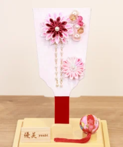 Handcrafted Japanese Hagoita paddle in soft pastel pink tones, adorned with white and pink tsumami kanzashi flowers, pearl accents, and decorative beads. Displayed in a clear acrylic case with wooden base. By Yuubi. Wholesale by Fujiki Crafts.