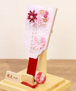 Handcrafted Japanese Hagoita paddle in soft pastel pink tones, adorned with white and pink tsumami kanzashi flowers, pearl accents, and decorative beads. Displayed in a clear acrylic case with wooden base. By Yuubi. Wholesale by Fujiki Crafts.