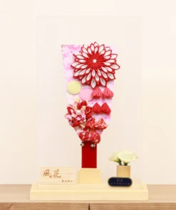 Traditional Japanese Hagoita paddle featuring vibrant red and white tsumami kanzashi flowers, sakura-patterned background, and hanging ornaments. Displayed in a clear acrylic case with wooden base, and decorative white rose accent. By Huka. Wholesale by Fujiki Crafts.