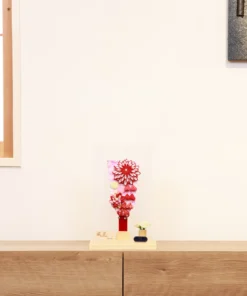 Traditional Japanese Hagoita paddle featuring vibrant red and white tsumami kanzashi flowers, sakura-patterned background, and hanging ornaments. Displayed in a clear acrylic case with wooden base, and decorative white rose accent. By Huka. Wholesale by Fujiki Crafts.