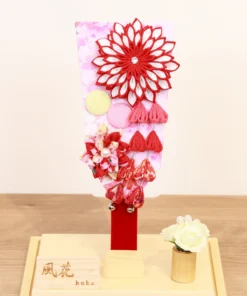 Traditional Japanese Hagoita paddle featuring vibrant red and white tsumami kanzashi flowers, sakura-patterned background, and hanging ornaments. Displayed in a clear acrylic case with wooden base, and decorative white rose accent. By Huka. Wholesale by Fujiki Crafts.