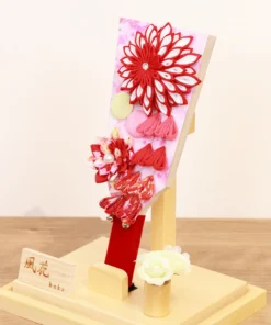 Traditional Japanese Hagoita paddle featuring vibrant red and white tsumami kanzashi flowers, sakura-patterned background, and hanging ornaments. Displayed in a clear acrylic case with wooden base, and decorative white rose accent. By Huka. Wholesale by Fujiki Crafts.