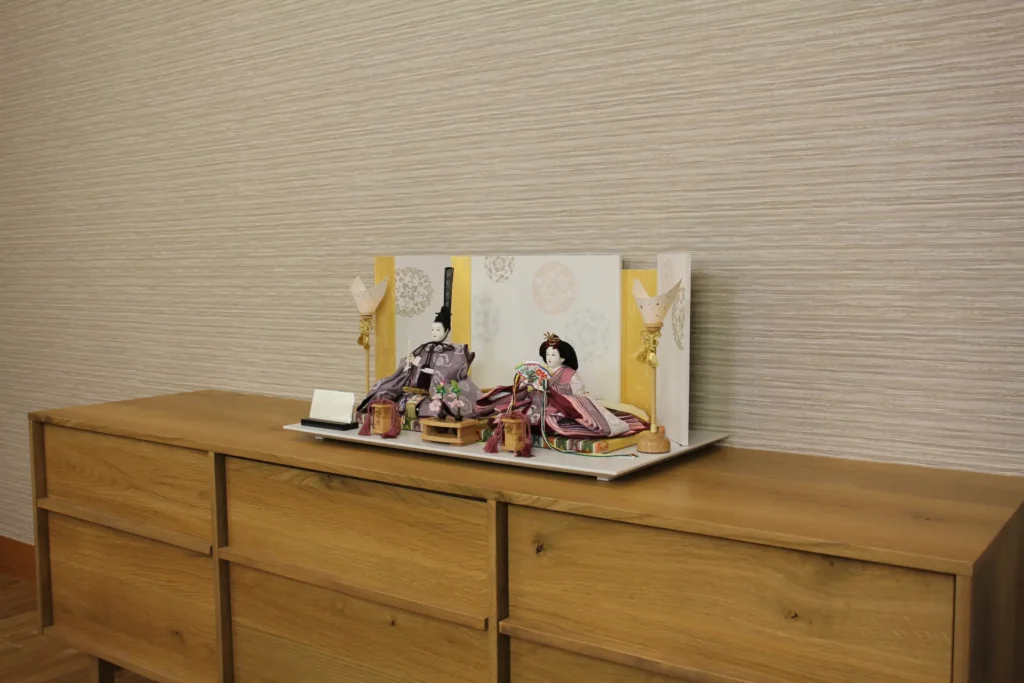 Elegant Japanese Hina doll set with plum and burgundy kimono, displayed on a white platform with gold-accented folding screen and lanterns. Wholesale by Fujiki Crafts.