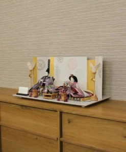 Elegant Japanese Hina doll set with plum and burgundy kimono, displayed on a white platform with gold-accented folding screen and lanterns. Wholesale by Fujiki Crafts.