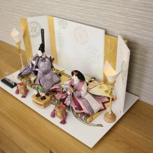 Elegant Japanese Hina doll set with plum and burgundy kimono, displayed on a white platform with gold-accented folding screen and lanterns. Wholesale by Fujiki Crafts.