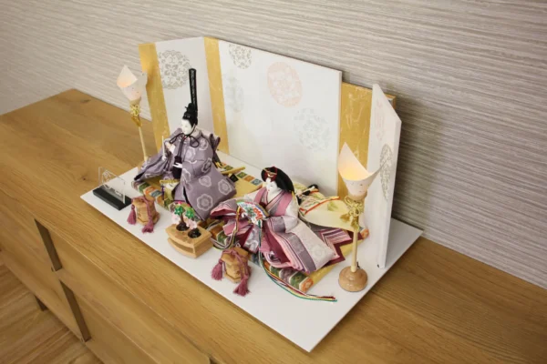 Elegant Japanese Hina doll set with plum and burgundy kimono, displayed on a white platform with gold-accented folding screen and lanterns. Wholesale by Fujiki Crafts.