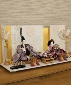 Elegant Japanese Hina doll set with plum and burgundy kimono, displayed on a white platform with gold-accented folding screen and lanterns. Wholesale by Fujiki Crafts.