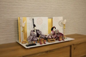Elegant Japanese Hina doll set with plum and burgundy kimono, displayed on a white platform with gold-accented folding screen and lanterns. Wholesale by Fujiki Crafts.