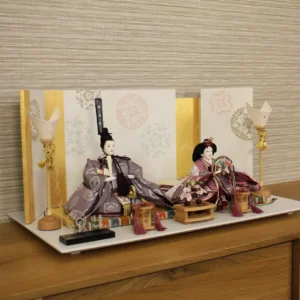 Elegant Japanese Hina doll set with plum and burgundy kimono, displayed on a white platform with gold-accented folding screen and lanterns. Wholesale by Fujiki Crafts.