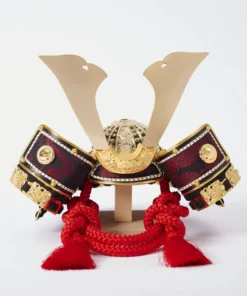 Handcrafted Japanese kabuto samurai helmet display with red cord and gold accents, set on a wooden stand with katana and arrows. Wholesale by Fujiki Crafts.