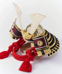 Handcrafted Japanese kabuto samurai helmet display with red cord and gold accents, set on a wooden stand with katana and arrows. Wholesale by Fujiki Crafts.
