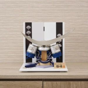 Japanese samurai kabuto helmet in silver with blue cords and tassels, mounted on a white and wood display stand with decorative folding screen. Wholesale by Fujiki Crafts.