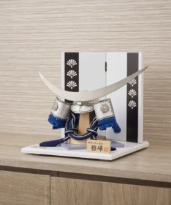 Japanese samurai kabuto helmet in silver with blue cords and tassels, mounted on a white and wood display stand with decorative folding screen. Wholesale by Fujiki Crafts.