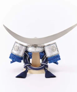 Japanese samurai kabuto helmet in silver with blue cords and tassels, mounted on a white and wood display stand with decorative folding screen. Wholesale by Fujiki Crafts.