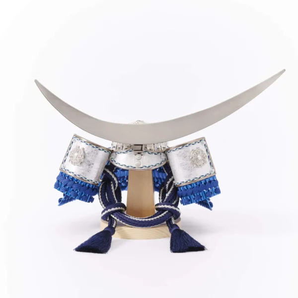 Japanese samurai kabuto helmet in silver with blue cords and tassels, mounted on a white and wood display stand with decorative folding screen. Wholesale by Fujiki Crafts.