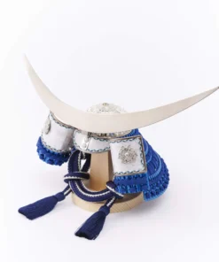 Japanese samurai kabuto helmet in silver with blue cords and tassels, mounted on a white and wood display stand with decorative folding screen. Wholesale by Fujiki Crafts.