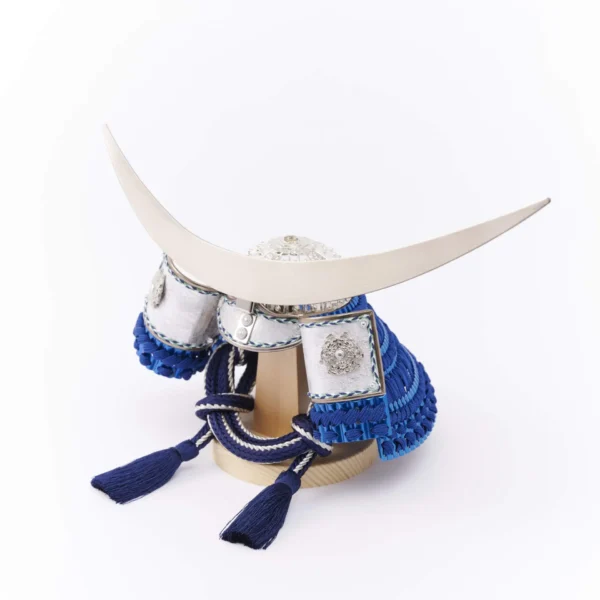 Japanese samurai kabuto helmet in silver with blue cords and tassels, mounted on a white and wood display stand with decorative folding screen. Wholesale by Fujiki Crafts.