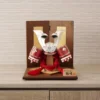 Japanese Kabuto samurai helmet with bold red cords and gold detailing, displayed on a wooden base with folding screen backdrop. Wholesale by Fujiki Crafts.