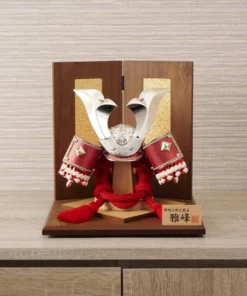 Japanese Kabuto samurai helmet with bold red cords and gold detailing, displayed on a wooden base with folding screen backdrop. Wholesale by Fujiki Crafts.